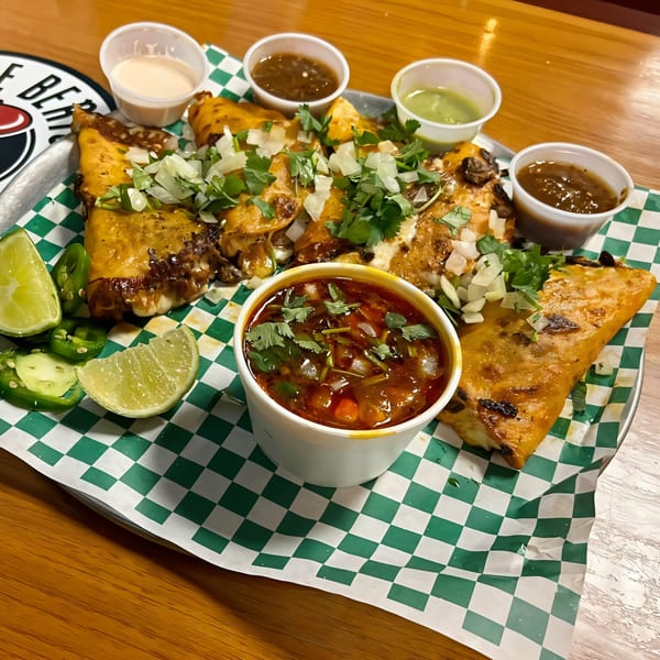 Uncle Berto's Burritos birria tacos with consome, limes, jalapeños, and various sauces on a tray.