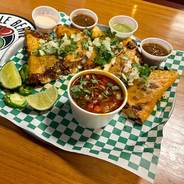A tray of cheesy birria tacos with cilantro, onions, lime, jalapeños, and dipping sauces.