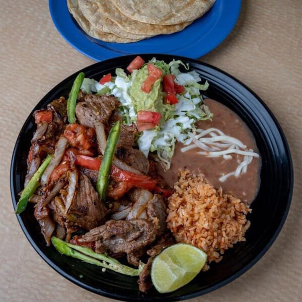 A black plate of carne asada with rice, refried beans, guacamole, and a lime wedge.