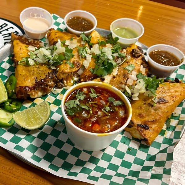 Uncle Berto's Burritos birria tacos with consome, limes, and salsas on a checkered paper-lined tray.